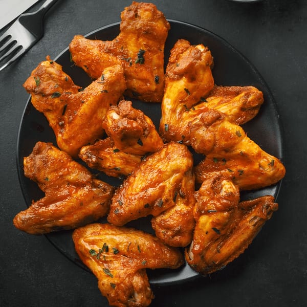 A black plate filled with golden-brown Angelo's Pizza & Subs chicken wings, coated in sauce and herbs.