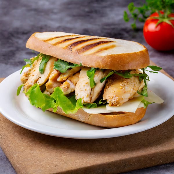 Grilled chicken sub with lettuce, cheese, and arugula on a white plate with a tomato.